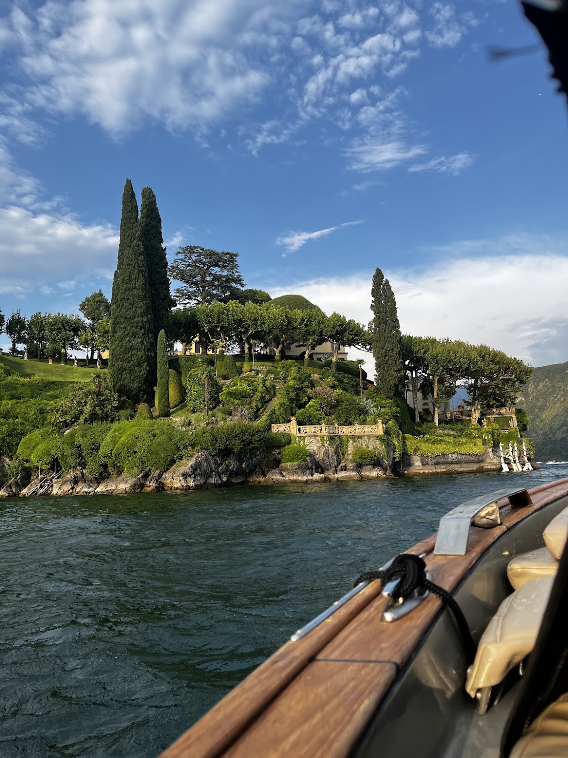 Terraced villa gardens with tall cypresses and a stone balustrade on Lake Como, seen from the timber rail of the boat