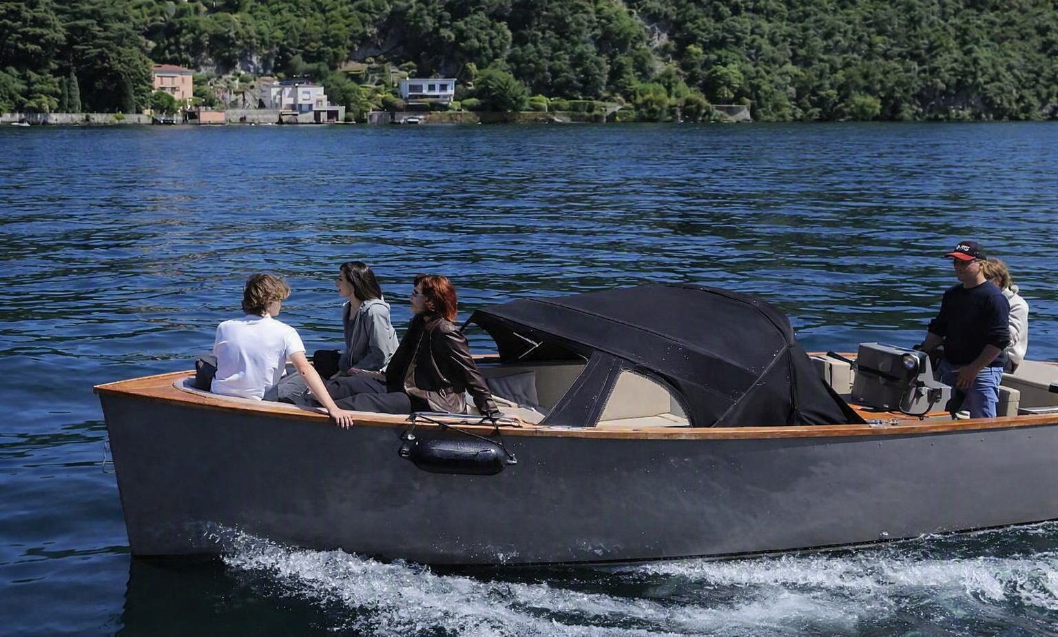The Cooper 800 under way on Lake Como, four guests seated in the open cockpit with the skipper at the helm and a green wooded shoreline with villas behind