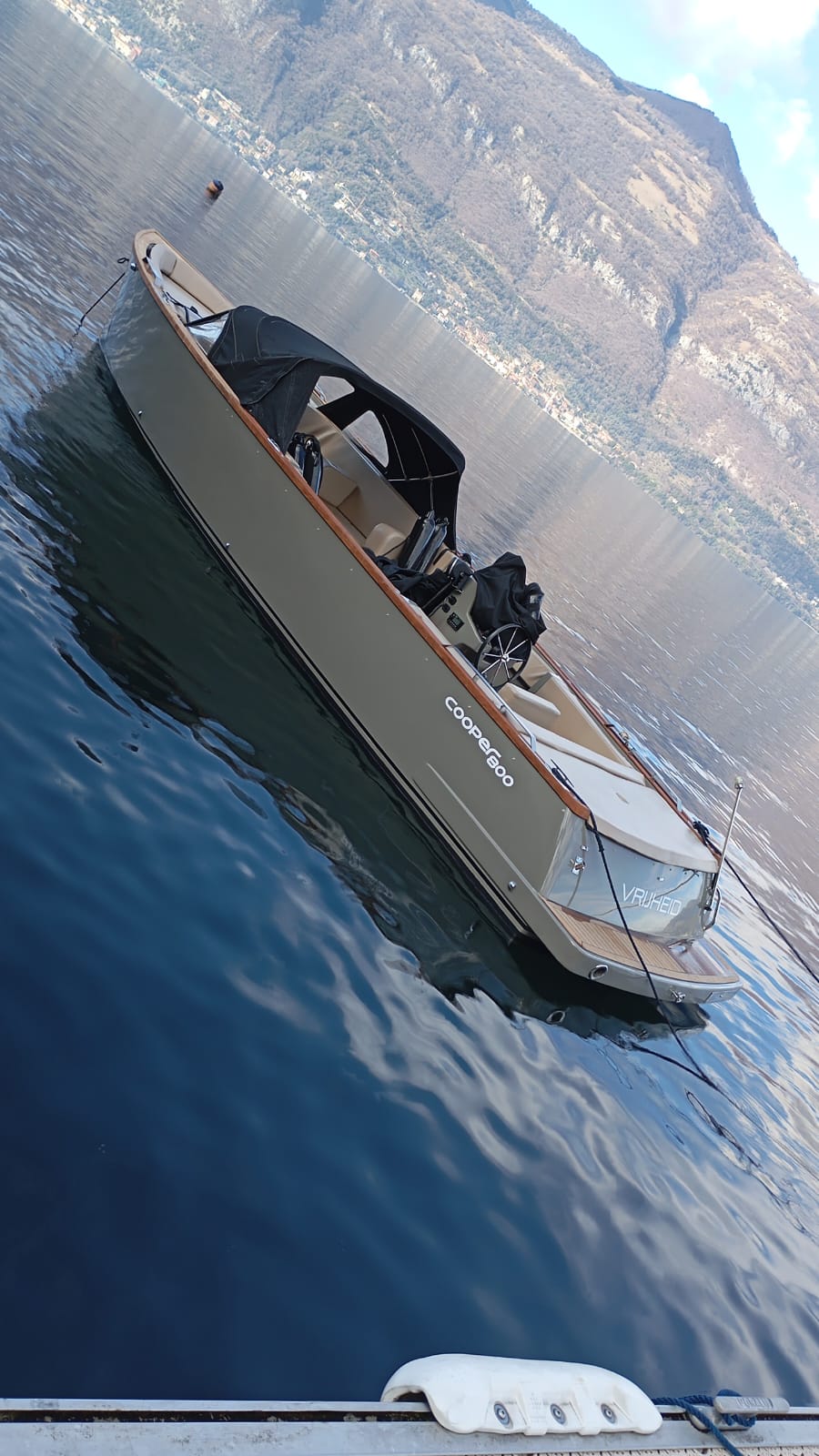 Real side profile of the grey-green Cooper 800 moored on Lake Como, showing its timber gunwale, black bimini, open helm and cream seating